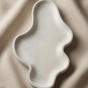 May include: An off-white tray with an irregular, organic, wavy shape and a subtle marbled texture. The surface is smooth and features soft, light gray swirls throughout the material, creating a distinct pattern within the curved edges.