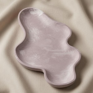 May include: An irregular, organic-shaped tray featuring a smooth, matte finish in a soft, muted lavender color with subtle, swirling white marbled patterns throughout the surface, creating a unique, flowing silhouette with raised, rounded edges.