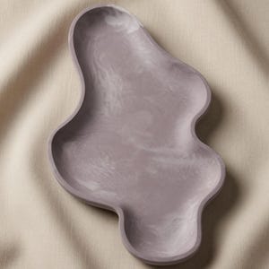 May include: A lavender-colored tray with an irregular, organic, and wavy shape. The surface features a subtle, marbled texture with lighter and darker shades of purple blended throughout the smooth, matte finish of the shallow dish.