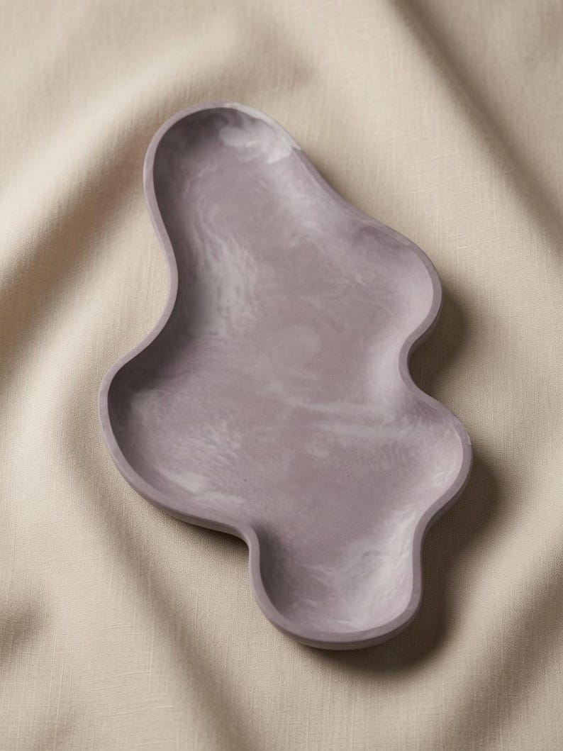 May include: A lavender-colored tray with an irregular, organic, and wavy shape. The surface features a subtle, marbled texture with lighter and darker shades of purple blended throughout the smooth, matte finish of the shallow dish.