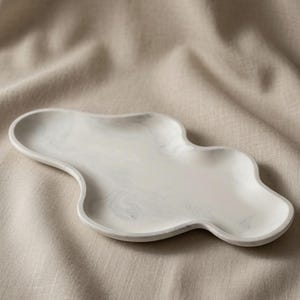 May include: An oblong, cloud-shaped tray featuring a white base with subtle, swirling gray marble-like patterns. The tray has a smooth, raised edge and an irregular, organic perimeter, presenting a flat, shallow surface for holding small items.