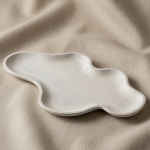 May include: An irregular, cloud-shaped tray featuring a smooth, off-white surface with subtle, swirling gray marble-like patterns. The tray has a shallow, concave center and a raised, rounded rim, presenting a continuous, fluid, and organic form.