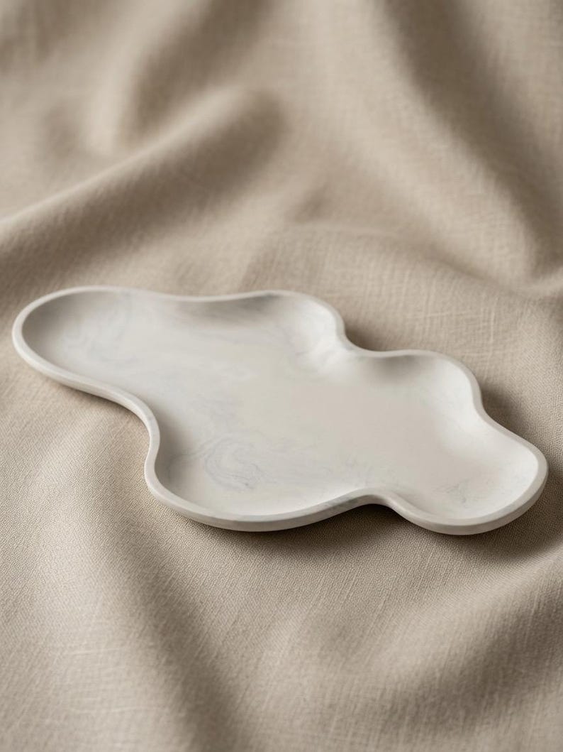 May include: An irregular, cloud-shaped tray featuring a smooth, off-white surface with subtle, swirling gray marble-like patterns. The tray has a shallow, concave center and a raised, rounded rim, presenting a continuous, fluid, and organic form.