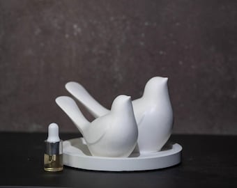 May include: Two smooth white ceramic bird figurines are positioned on a matching circular white tray. Next to the tray stands a small clear glass bottle with a white dropper cap containing a light amber liquid, completing the set.
