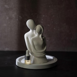 Puede incluir: Una escultura blanca mate de dos figuras abrazadas, colocada sobre una bandeja circular blanca. Un pequeño frasco de vidrio con tapa de gotero plateada que contiene un líquido transparente se encuentra en la bandeja frente a las figuras.