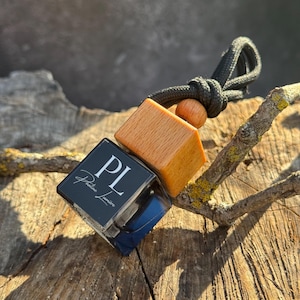 May include: A square glass car air freshener bottle with a black cap and a light wood topper. The front of the glass features a black label with the white letters PL and the text Pauline Lumière. A black cord is attached to the wooden topper.