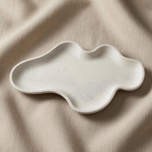 May include: An off-white, cloud-shaped tray with a smooth, matte finish and subtle gray marbling throughout the surface. The tray features an irregular, organic perimeter with rounded edges and a shallow, concave center for holding small items.