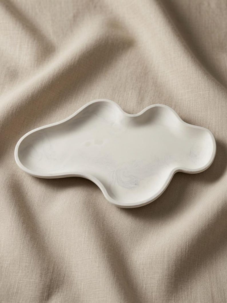May include: An off-white, cloud-shaped tray with a smooth, matte finish and subtle gray marbling throughout the surface. The tray features an irregular, organic perimeter with rounded edges and a shallow, concave center for holding small items.