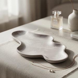 May include: A light gray, smooth, organic-shaped tray with a shallow basin and curved, undulating edges. The surface features a subtle, marbled texture in varying shades of gray, creating a soft, neutral appearance for this decorative object.