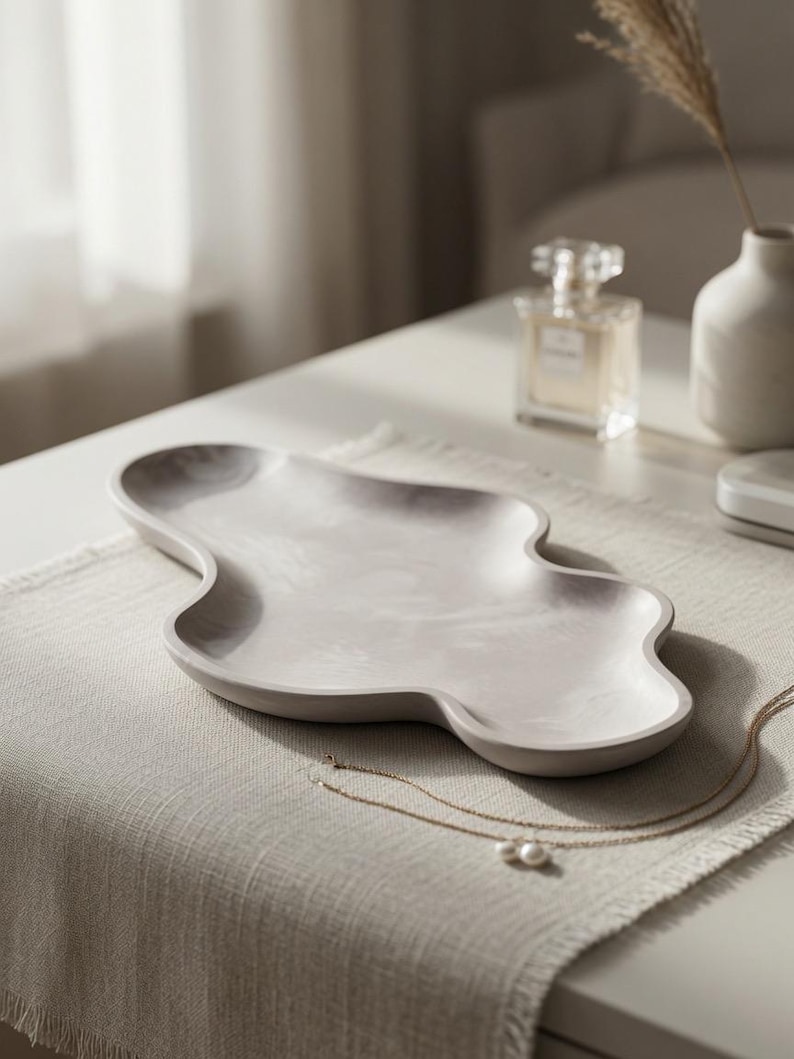 May include: A light gray, smooth, organic-shaped tray with a shallow basin and curved, undulating edges. The surface features a subtle, marbled texture in varying shades of gray, creating a soft, neutral appearance for this decorative object.