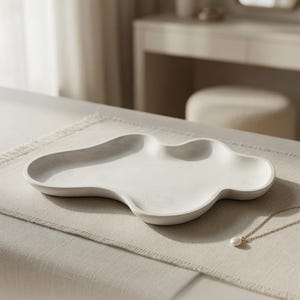 May include: An off-white, cloud-shaped decorative tray with a smooth, matte finish and raised edges. The tray measures 11.5 inches by 7.16 inches. A delicate gold chain necklace with a single white pearl pendant rests near the edge of the tray.