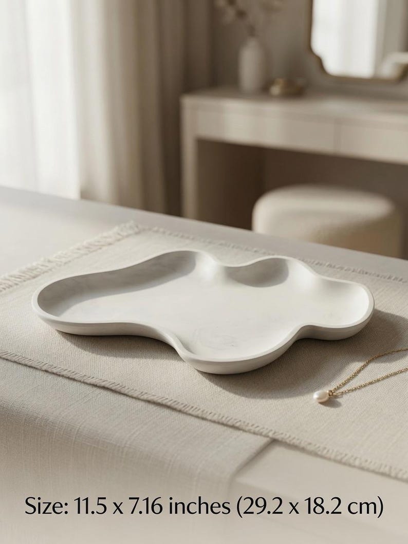 May include: An off-white, cloud-shaped decorative tray with a smooth, matte finish and raised edges. The tray measures 11.5 inches by 7.16 inches. A delicate gold chain necklace with a single white pearl pendant rests near the edge of the tray.