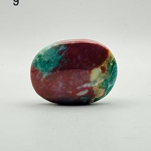 May include: A polished, oval-shaped stone featuring a vibrant mix of deep maroon, teal, and cream-coloured patterns. The surface is smooth and glossy, displaying natural mineral inclusions and colour variations throughout the entire piece.