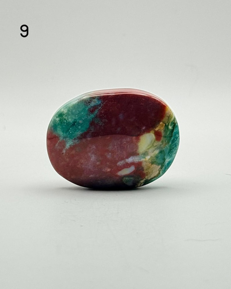 May include: A polished, oval-shaped stone featuring a vibrant mix of deep maroon, teal, and cream-coloured patterns. The surface is smooth and glossy, displaying natural mineral inclusions and colour variations throughout the entire piece.