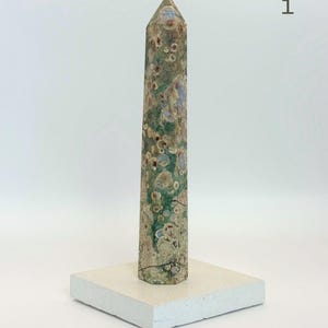 May include: A tall, hexagonal stone obelisk featuring a mottled pattern of green, tan, and brown circular inclusions. The stone tower stands vertically on a flat, square, off-white base. The number 1 is printed in the upper right corner.