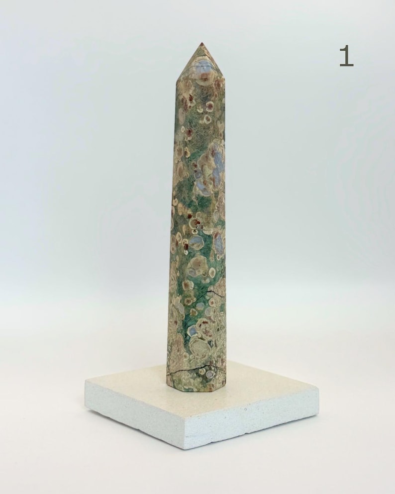 May include: A tall, hexagonal stone obelisk featuring a mottled pattern of green, tan, and brown circular inclusions. The stone tower stands vertically on a flat, square, off-white base. The number 1 is printed in the upper right corner.