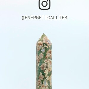 May include: A polished green crystal tower with circular tan and brown inclusions throughout. Above the crystal, the Instagram logo is displayed alongside the text @ENERGETICALLIES. The stone features a pointed top and a cylindrical shape.