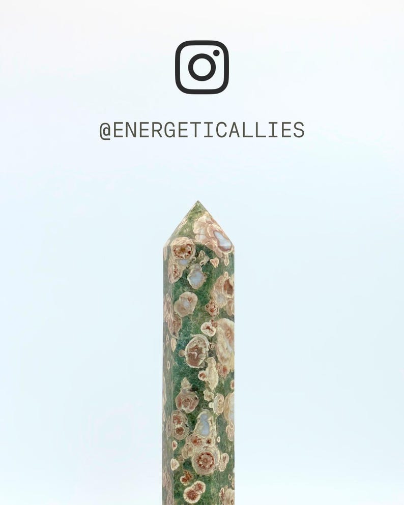 May include: A polished green crystal tower with circular tan and brown inclusions throughout. Above the crystal, the Instagram logo is displayed alongside the text @ENERGETICALLIES. The stone features a pointed top and a cylindrical shape.