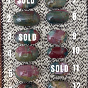 May include: Twelve oval-shaped polished bloodstone cabochons arranged in two columns, numbered one through twelve. Each stone features a unique blend of deep green, red, and brown hues. The stones labelled one, three, and eleven have the word SOLD.