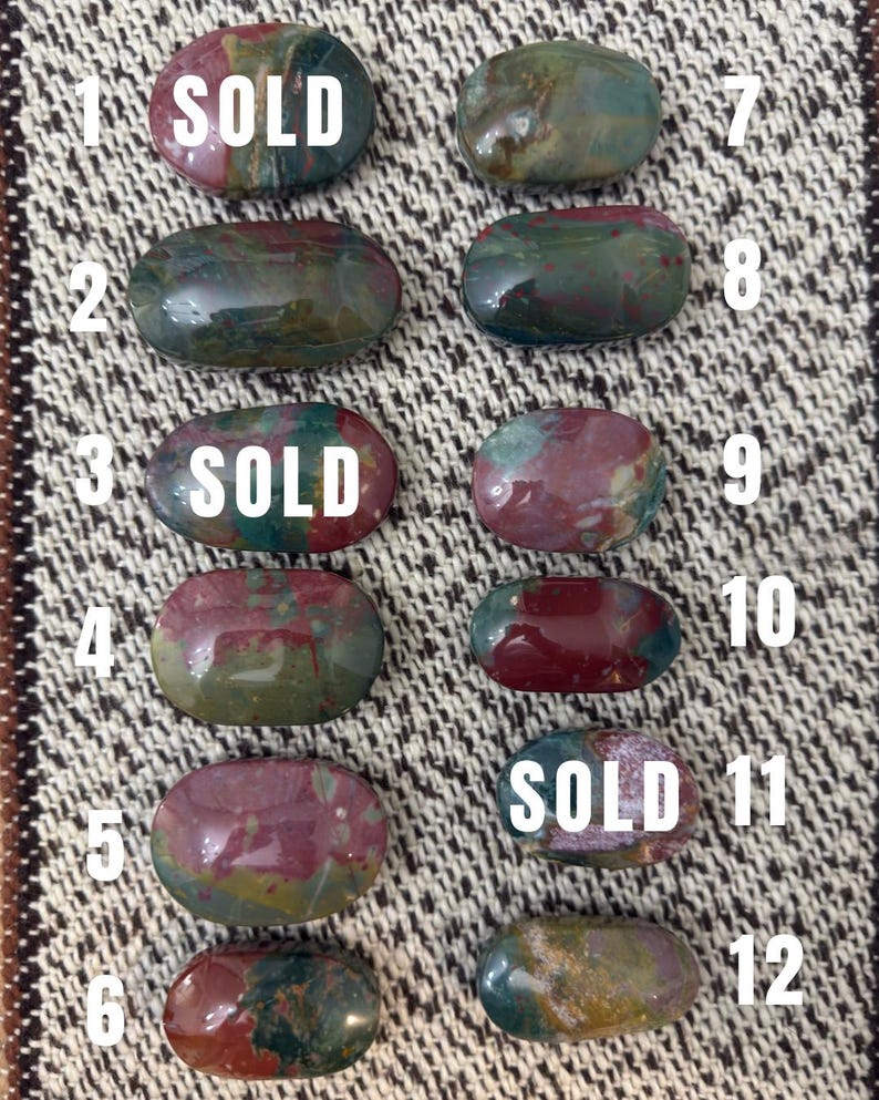 May include: Twelve oval-shaped polished bloodstone cabochons arranged in two columns, numbered one through twelve. Each stone features a unique blend of deep green, red, and brown hues. The stones labelled one, three, and eleven have the word SOLD.