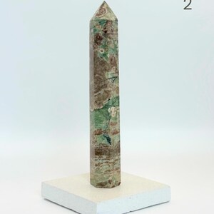 May include: A tall, hexagonal stone obelisk featuring a mottled pattern of sage green, earthy brown, and cream-colored inclusions. The polished mineral tower stands upright on a square, off-white base. The number 2 is visible in the top right corner.