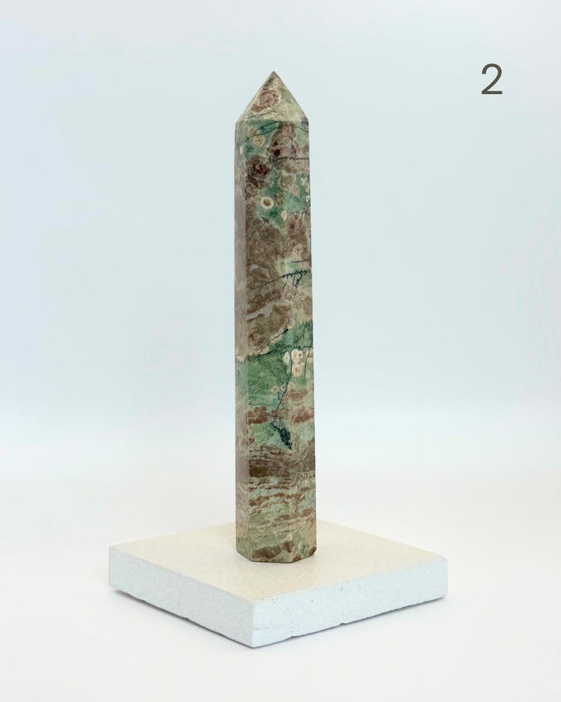 May include: A tall, hexagonal stone obelisk featuring a mottled pattern of sage green, earthy brown, and cream-colored inclusions. The polished mineral tower stands upright on a square, off-white base. The number 2 is visible in the top right corner.