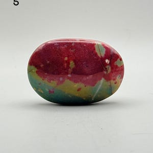 May include: An oval-shaped polished stone featuring a vibrant colour palette of deep magenta, muted olive green, and soft teal. The surface is smooth and glossy, displaying natural mineral inclusions and subtle white veining throughout the piece.