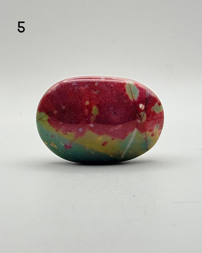 May include: An oval-shaped polished stone featuring a vibrant colour palette of deep magenta, muted olive green, and soft teal. The surface is smooth and glossy, displaying natural mineral inclusions and subtle white veining throughout the piece.