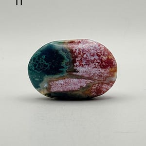 May include: A polished, oval-shaped stone featuring a vibrant mix of deep forest green, bright crimson red, and creamy white patterns. The surface displays intricate, swirling natural inclusions and a smooth, glossy finish throughout the piece.