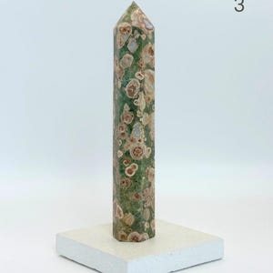 May include: A tall, hexagonal crystal tower featuring a mottled green base with circular, flower-like patterns in shades of tan, brown, and cream. The stone point stands upright on a flat, square, off-white base. The number 3 is visible.