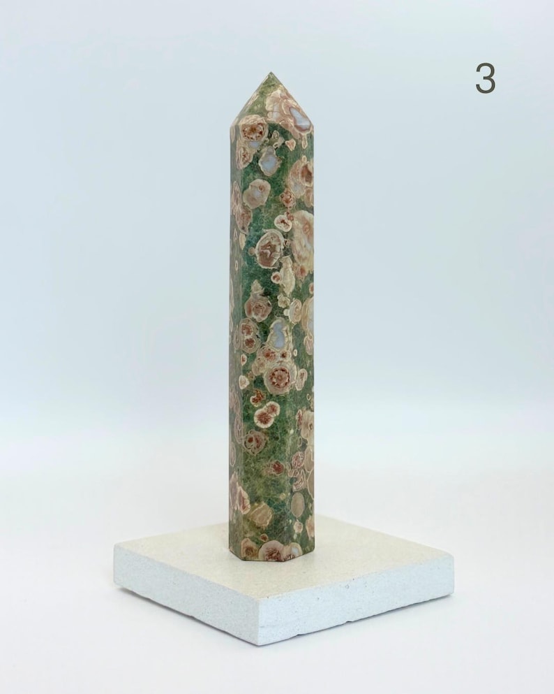 May include: A tall, hexagonal crystal tower featuring a mottled green base with circular, flower-like patterns in shades of tan, brown, and cream. The stone point stands upright on a flat, square, off-white base. The number 3 is visible.