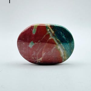 May include: A polished, oval-shaped stone featuring a smooth surface with distinct colour zones. The stone displays a deep red section on the left, transitioning into a vibrant teal and green area on the right, separated by thin white veins.