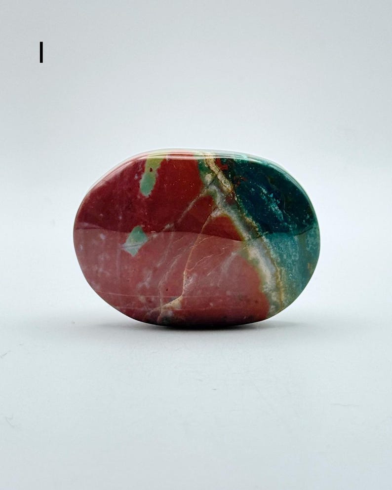 May include: A polished, oval-shaped stone featuring a smooth surface with distinct colour zones. The stone displays a deep red section on the left, transitioning into a vibrant teal and green area on the right, separated by thin white veins.