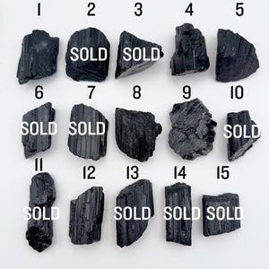 May include: A collection of fifteen raw black tourmaline crystals arranged in a grid, each labeled with a number from 1 to 15. Several of the crystals are marked with the word SOLD in white text. The stones feature natural vertical striations.