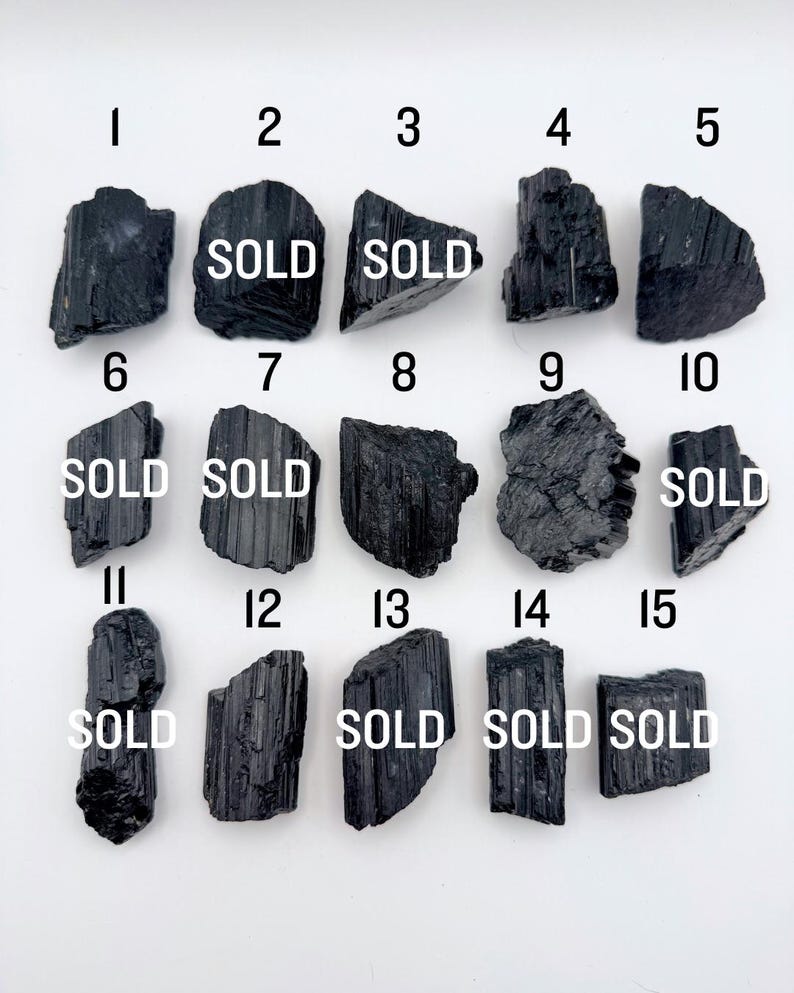 May include: A collection of fifteen raw black tourmaline crystals arranged in a grid, each labeled with a number from 1 to 15. Several of the crystals are marked with the word SOLD in white text. The stones feature natural vertical striations.