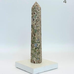 May include: A tall, six-sided obelisk carved from mottled green and brown stone with circular inclusions, standing upright on a flat, square, off-white stone base. The number 4 is visible in the upper right corner of the image.