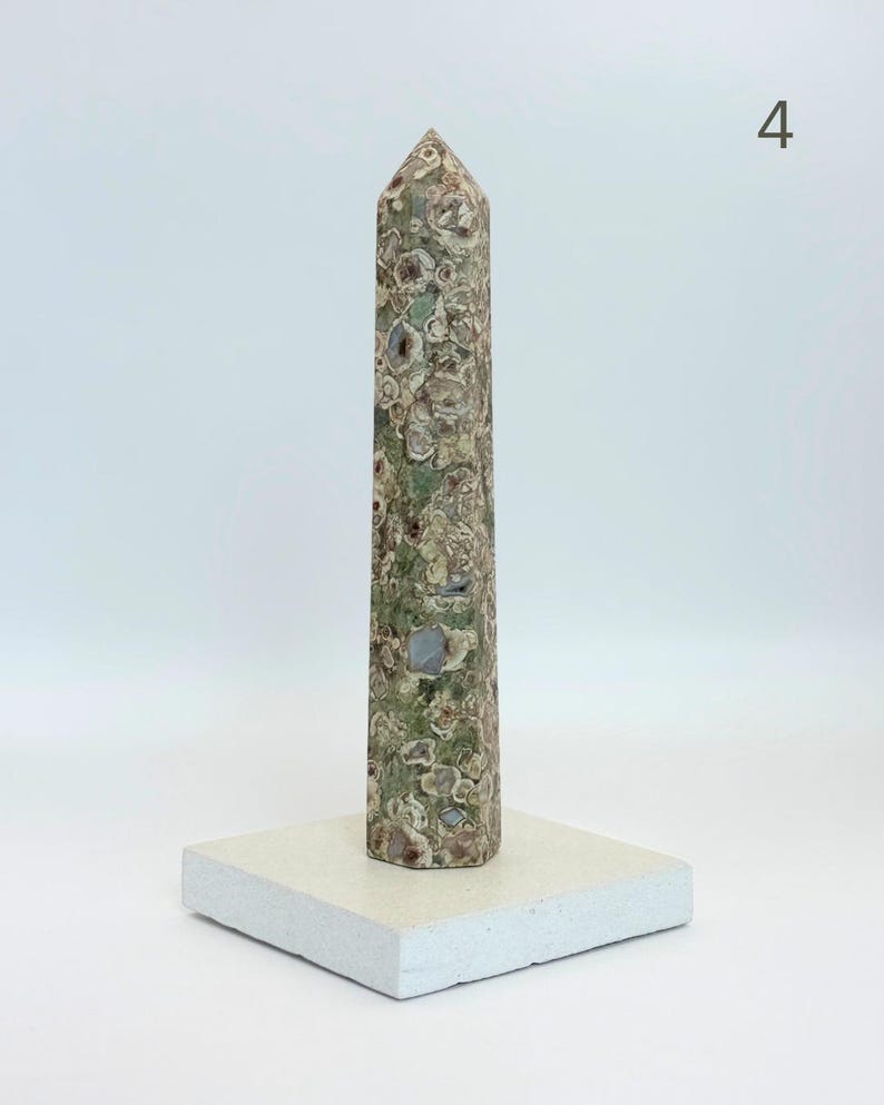 May include: A tall, six-sided obelisk carved from mottled green and brown stone with circular inclusions, standing upright on a flat, square, off-white stone base. The number 4 is visible in the upper right corner of the image.