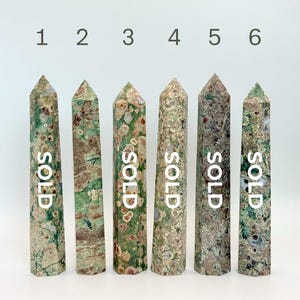 May include: Six tall, pointed crystal towers arranged in a row, numbered one through six. Each stone features a unique mottled pattern of green, brown, and cream colors. The text "SOLD" is printed vertically on five of the six crystal towers.