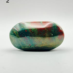May include: A polished, oval-shaped bloodstone specimen featuring a deep forest green base colour with prominent, irregular patches of vibrant red and subtle hints of yellow and translucent grey throughout the smooth, reflective surface.
