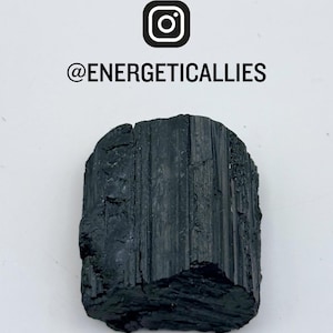 May include: A raw black tourmaline crystal with vertical striations and a rough, uneven texture. Above the crystal, the text reads @ENERGETICALLIES with an Instagram icon. The crystal has a dark, opaque appearance with natural fractures.