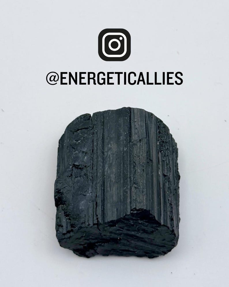 May include: A raw black tourmaline crystal with vertical striations and a rough, uneven texture. Above the crystal, the text reads @ENERGETICALLIES with an Instagram icon. The crystal has a dark, opaque appearance with natural fractures.