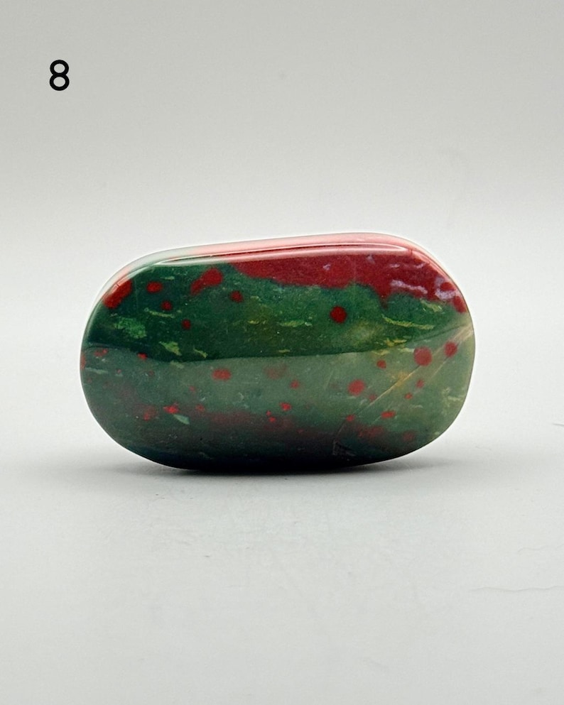 May include: A polished, oval-shaped bloodstone specimen featuring a deep forest green base colour with distinct, scattered bright red spots and patches throughout the surface. The stone has a smooth, glossy texture and is labelled with the number 8.