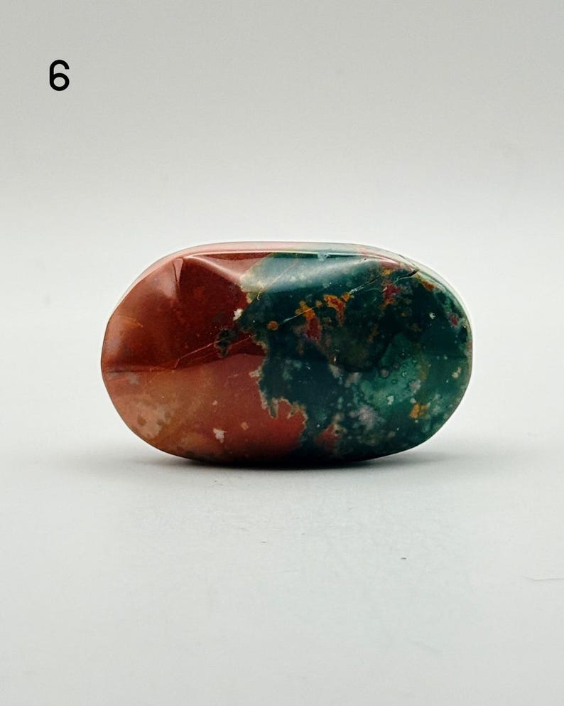 May include: A polished, oval-shaped stone featuring a distinct colour split between deep red and dark forest green. The surface is smooth and glossy, displaying natural mineral inclusions and subtle variations in tone across the entire rock.
