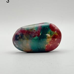 May include: A smooth, polished, oval-shaped stone featuring a vibrant, multi-coloured pattern with swirls of deep red, forest green, teal, and hints of yellow and white. The surface has a glossy, reflective finish that highlights the colour variations.