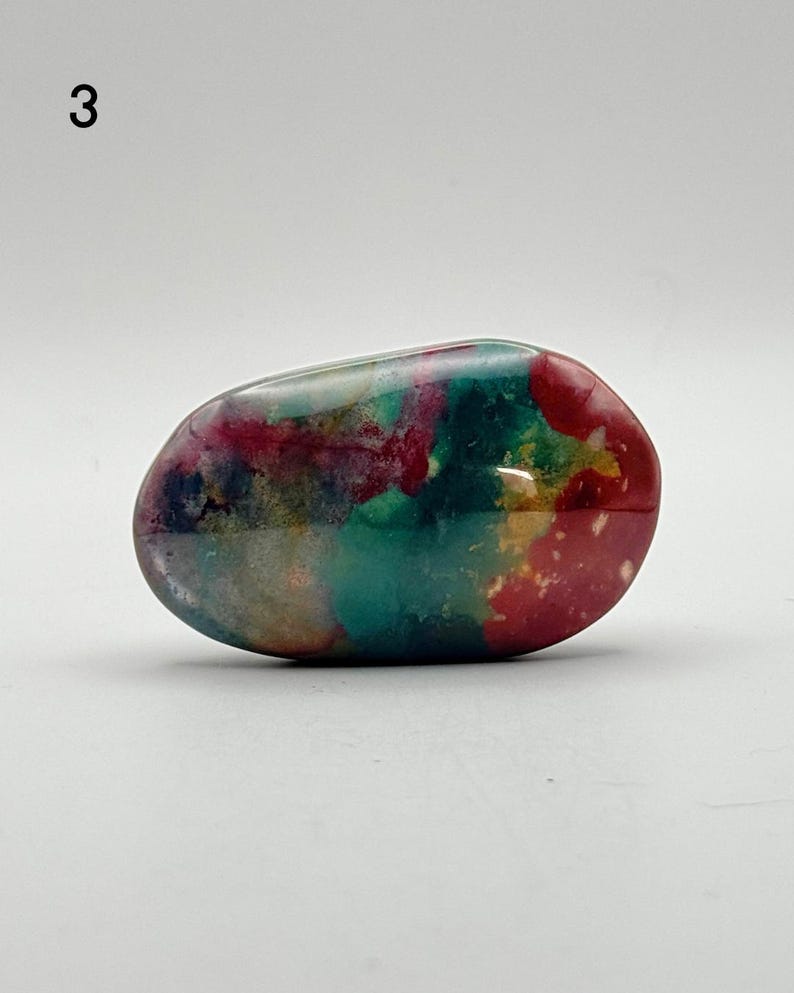 May include: A smooth, polished, oval-shaped stone featuring a vibrant, multi-coloured pattern with swirls of deep red, forest green, teal, and hints of yellow and white. The surface has a glossy, reflective finish that highlights the colour variations.
