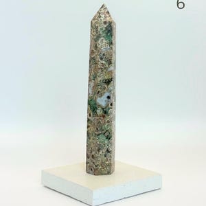 May include: A tall, multi-faceted obelisk carved from patterned stone featuring a complex mix of moss green, brown, tan, and cream circular inclusions. The obelisk stands vertically on a flat, square, off-white base. The number 6 is visible.