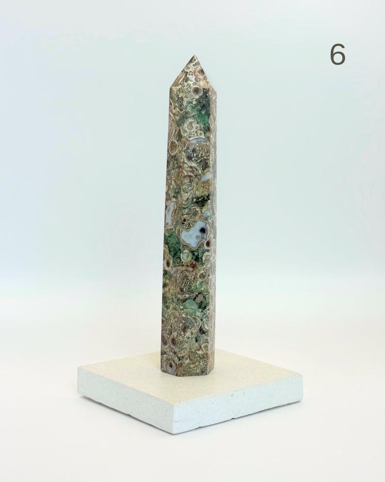 May include: A tall, multi-faceted obelisk carved from patterned stone featuring a complex mix of moss green, brown, tan, and cream circular inclusions. The obelisk stands vertically on a flat, square, off-white base. The number 6 is visible.