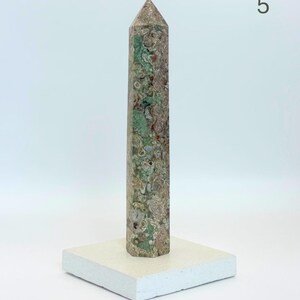 May include: A tall, hexagonal stone obelisk featuring a mottled pattern of sage green, earthy brown, and cream circular inclusions. The stone tower is mounted vertically on a flat, square, off-white base. The number 5 is visible in the top right corner.