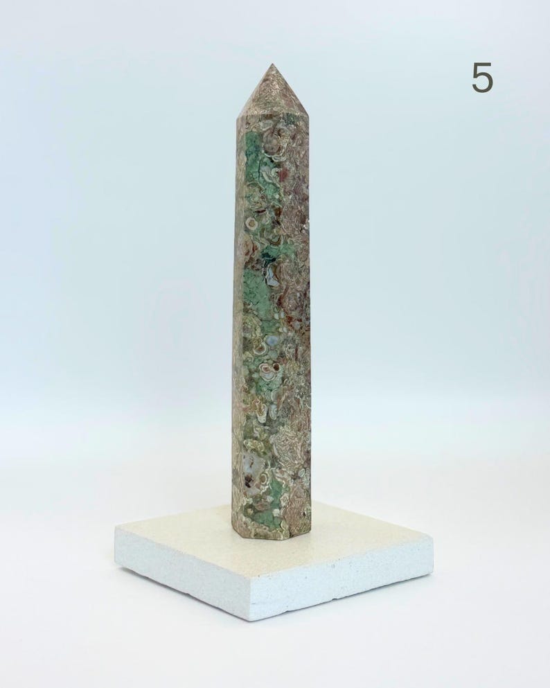 May include: A tall, hexagonal stone obelisk featuring a mottled pattern of sage green, earthy brown, and cream circular inclusions. The stone tower is mounted vertically on a flat, square, off-white base. The number 5 is visible in the top right corner.