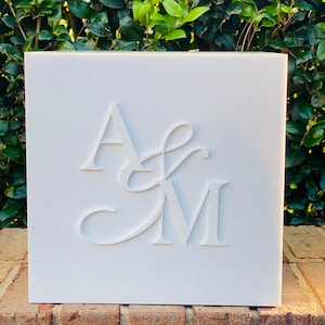 May include: A white square sign with the raised initials "A & M" in a decorative font. The sign is set against a backdrop of green foliage and a brick wall.